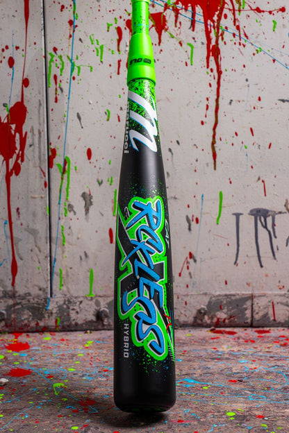A 2026 Marucci CAT X RCKLESS Hybrid (-5) USSSA baseball bat stands upright on a splattered paint floor, vibrant against a wall of colorful paint drips, highlighting its bold design and Marucci branding.