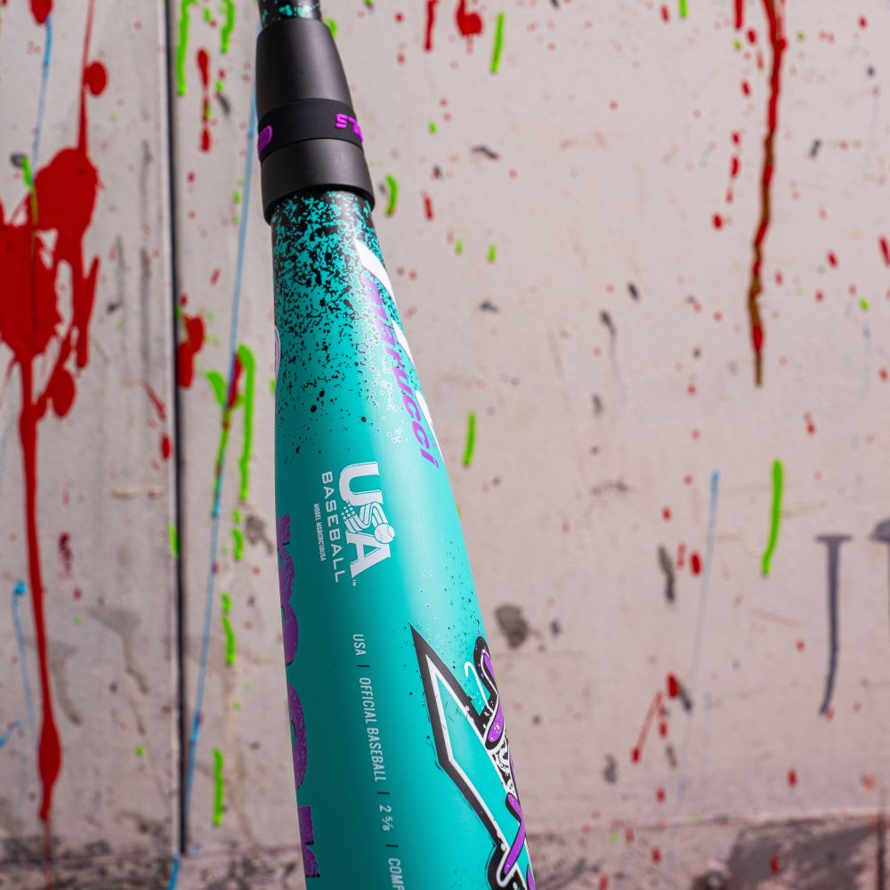 Close-up of the teal Marucci CAT X RCKLESS Composite (-10) 2 5/8" USA Baseball Bat (MSBCRC10USA) featuring a black handle and bold “USA Baseball” text, set against a white wall splattered with red, green, and blue paint.