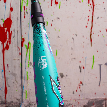 Close-up of the teal Marucci CAT X RCKLESS Composite (-10) 2 5/8" USA Baseball Bat (MSBCRC10USA) featuring a black handle and bold “USA Baseball” text, set against a white wall splattered with red, green, and blue paint.