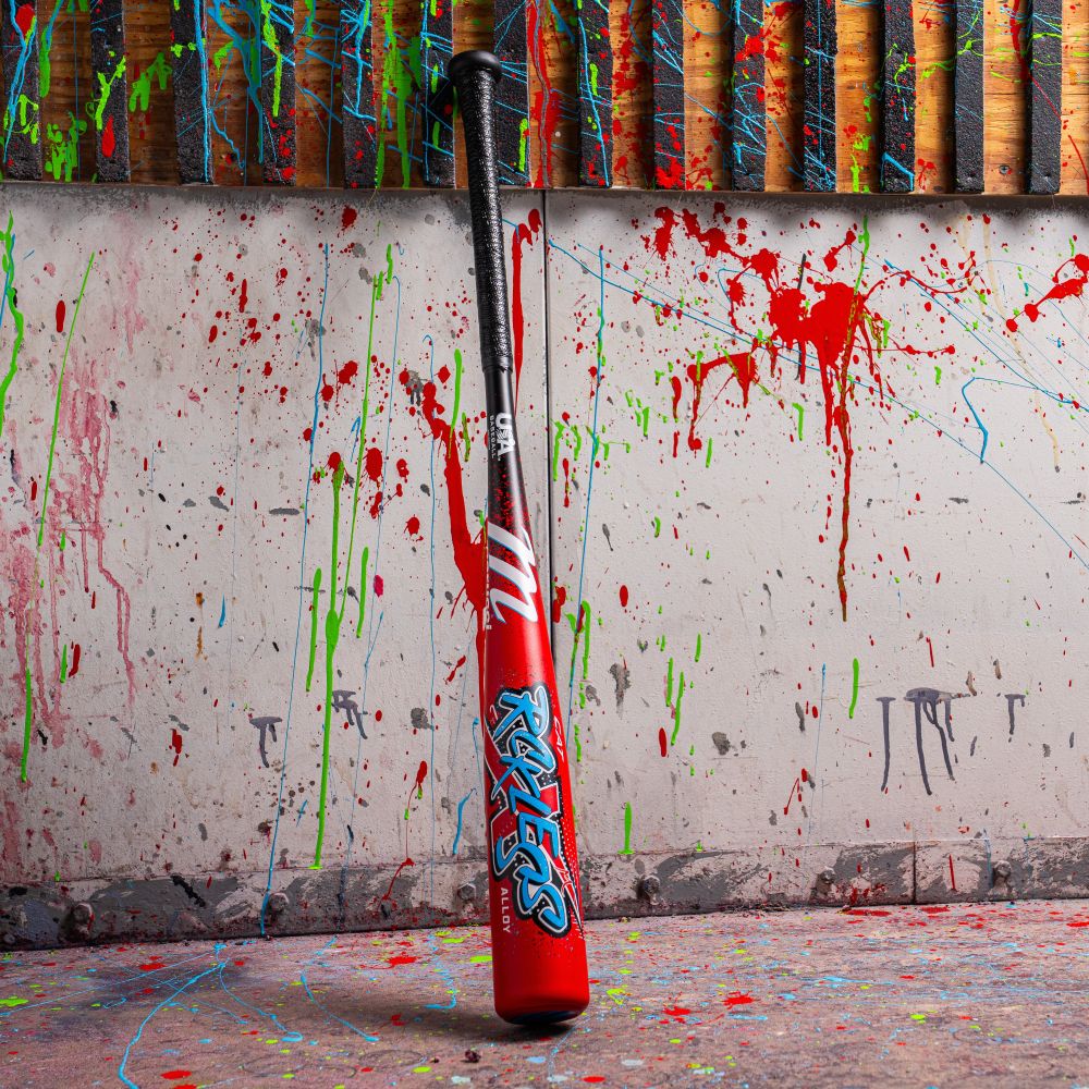 The Marucci 2026 CAT X RCKLESS (-8) USA Baseball Bat stands upright, its bold red and black design with blue and white “RITUAL” lettering contrasting against a wall and floor covered in vibrant paint splatters.
