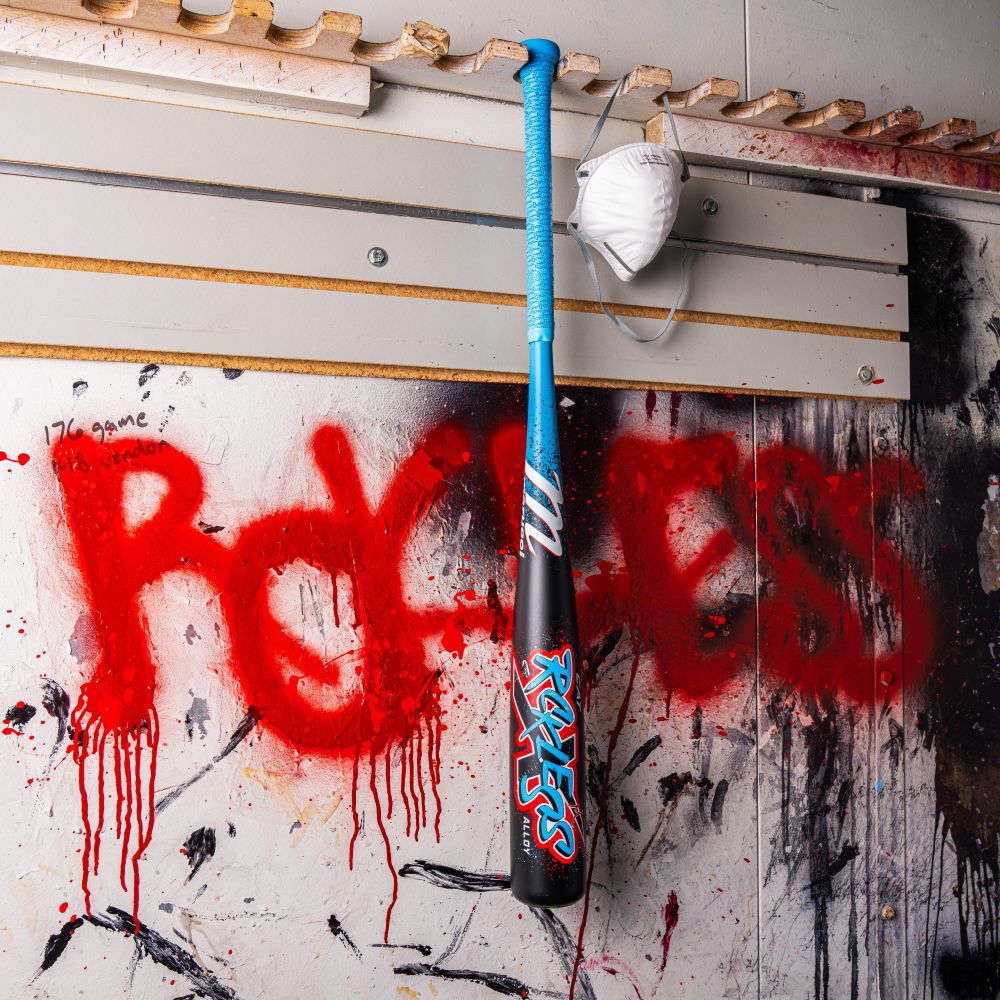 A Marucci 2026 CAT X RCKLESS (-8) USSSA Baseball Bat in blue and black hangs on a wall splattered with paint, next to a white face mask. "RAGELESS" is spray-painted in red on the wall behind them.