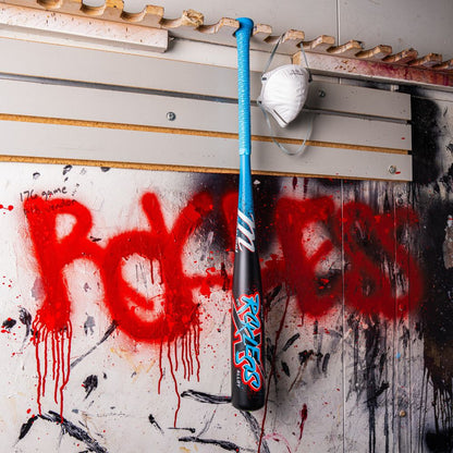 A Marucci 2026 CAT X RCKLESS (-8) USSSA Baseball Bat in blue and black hangs on a wall splattered with paint, next to a white face mask. "RAGELESS" is spray-painted in red on the wall behind them.