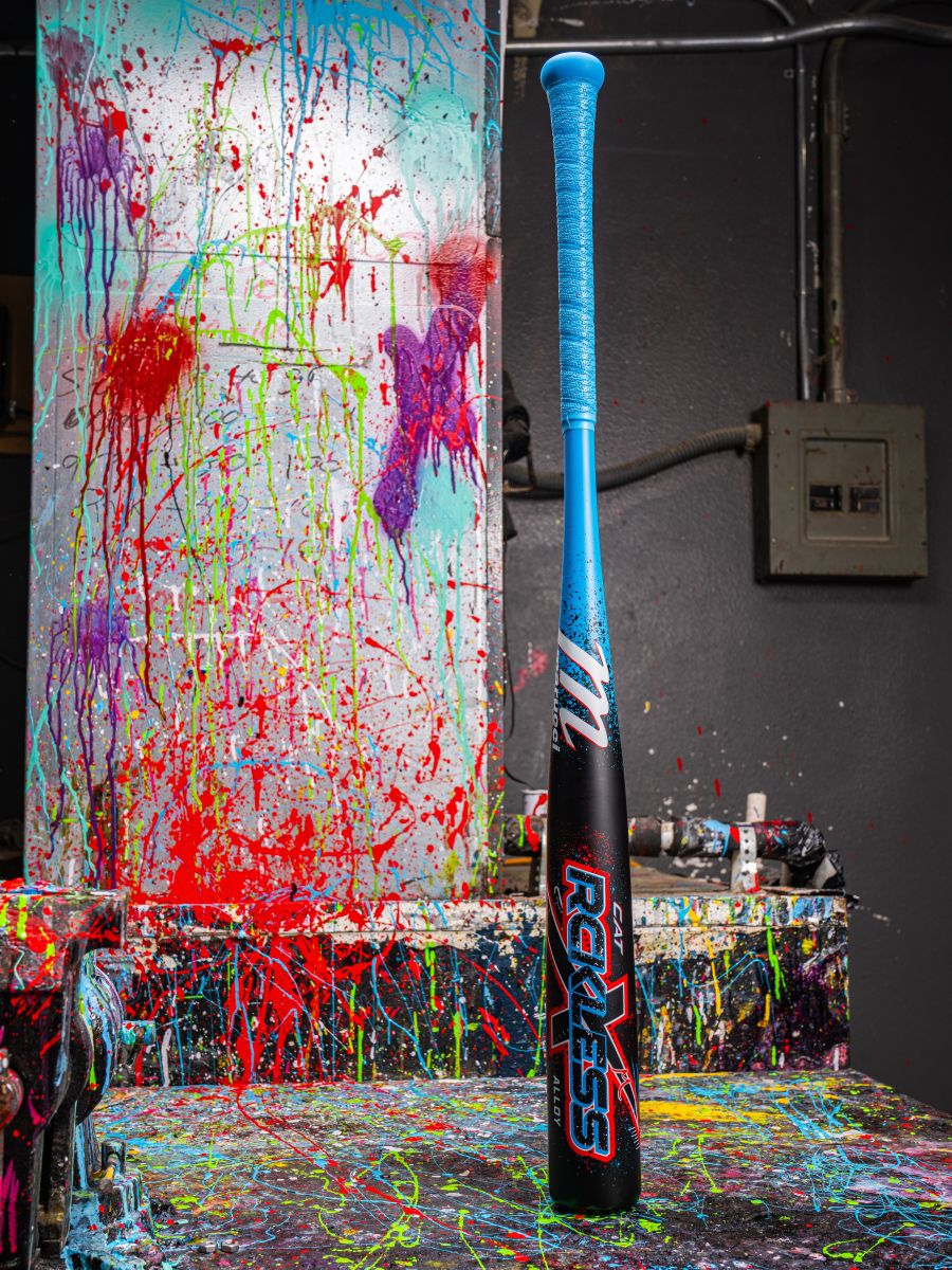 The Marucci 2026 CAT X RCKLESS (-3) BBCOR Baseball Bat stands upright on a paint-splattered table, with its blue and black design contrasting against a vibrant background of color-splashed walls and canvas.