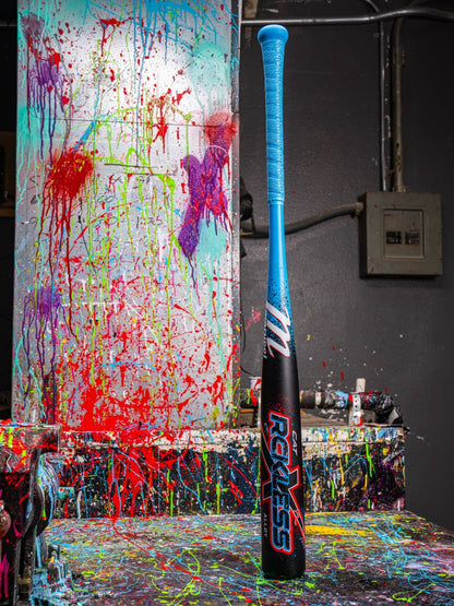 The Marucci 2026 CAT X RCKLESS (-3) BBCOR Baseball Bat stands upright on a paint-splattered table, with its blue and black design contrasting against a vibrant background of color-splashed walls and canvas.