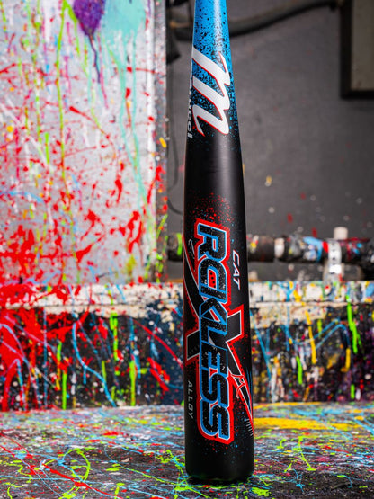 The Marucci 2026 CAT X RCKLESS (-3) BBCOR Baseball Bat stands upright with its bold graphics against a splattered, colorful background, vibrant paint drips and splashes covering the surrounding floor and wall.