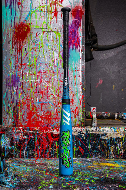 A blue Marucci 2026 CAT X RCKLESS Hybrid (-11) USA Baseball Bat (MSBCRH11USA) with graffiti-style graphics stands upright before a wall and floor covered in red, green, yellow, and pink paint splatters.