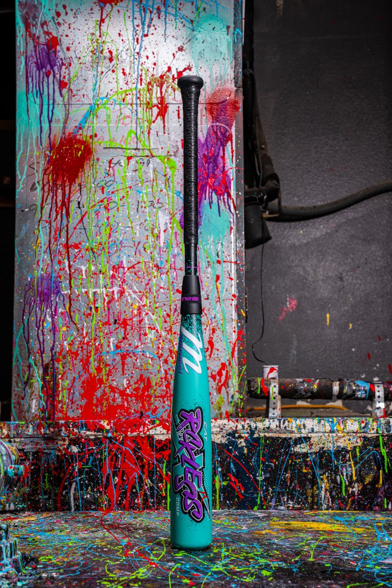 The Marucci 2026 CAT X RCKLESS Composite (-10) USA Baseball Bat stands upright, its turquoise barrel with graffiti-style graphics pops against a backdrop splattered with vibrant red, blue, yellow, and purple paint for a bold, artistic vibe.