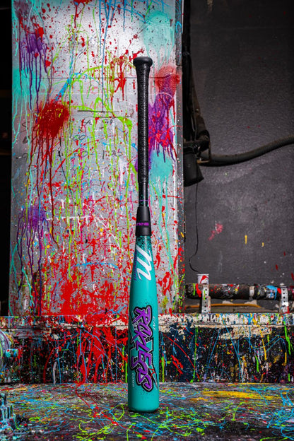 The Marucci 2026 CAT X RCKLESS Composite (-10) USA Baseball Bat stands upright, its turquoise barrel with graffiti-style graphics pops against a backdrop splattered with vibrant red, blue, yellow, and purple paint for a bold, artistic vibe.