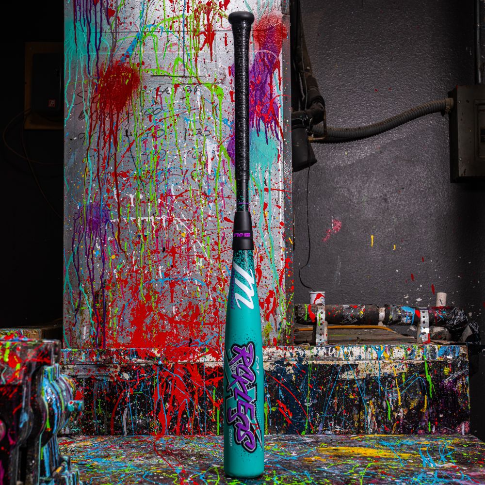 A vibrant Marucci 2026 CAT X RCKLESS Composite (-10) USA Baseball Bat stands upright on a splatter-painted table, bold and colorful against an industrial backdrop.
