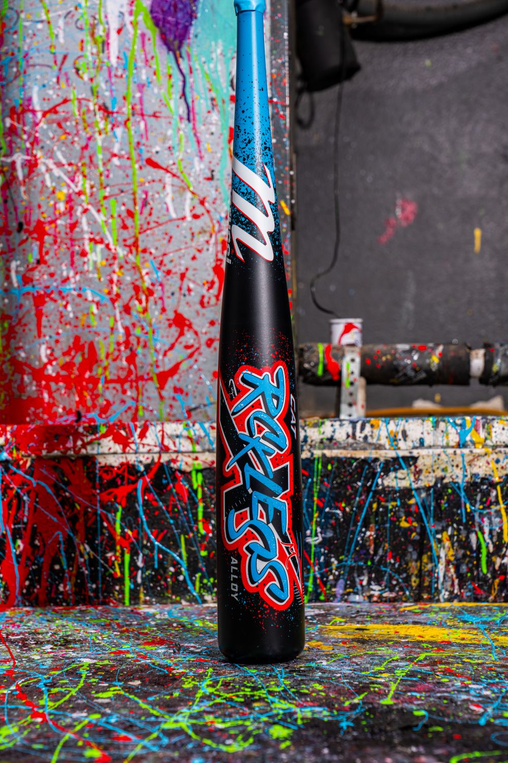 A black Marucci CAT X RCKLESS (-8) USSSA Baseball Bat (MSBCR8) stands upright against a paint-splattered wall, with splashes of blue, green, red, and yellow in the background and bold blue-red RCKLESS text on the barrel.