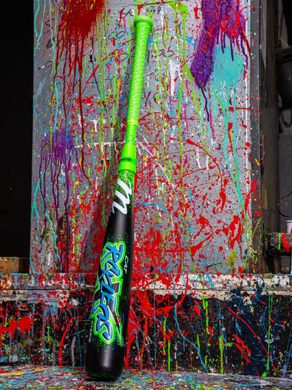 A Marucci 2026 CAT X RCKLESS Hybrid (-10) 2 3/4" USSSA Baseball Bat (MSBCRH10 DEMO) with black, green, and colorful paint splatters stands out against a concrete wall.