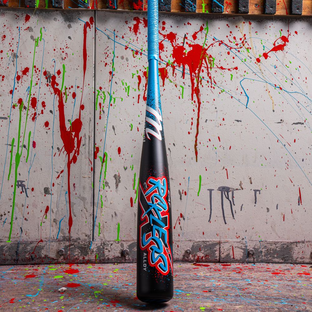 A 2026 Marucci CAT X RCKLESS (-8) USSSA Baseball Bat stands upright against a paint-splattered wall, featuring a blue handle and "Rockets" in red and blue letters, with colorful drips on the wall and floor.