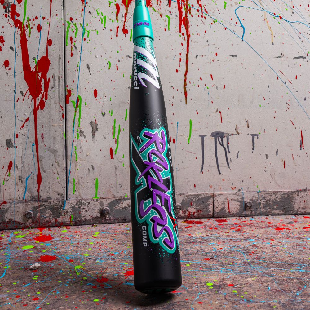 A black Marucci 2026 CAT X RCKLESS Composite (-5) USSSA baseball bat with neon graffiti-style "RULES" stands upright on a paint-splattered floor, echoing colorful splatters off the wall behind. This is the bold MSBCRC5 (DEMO) model in action.