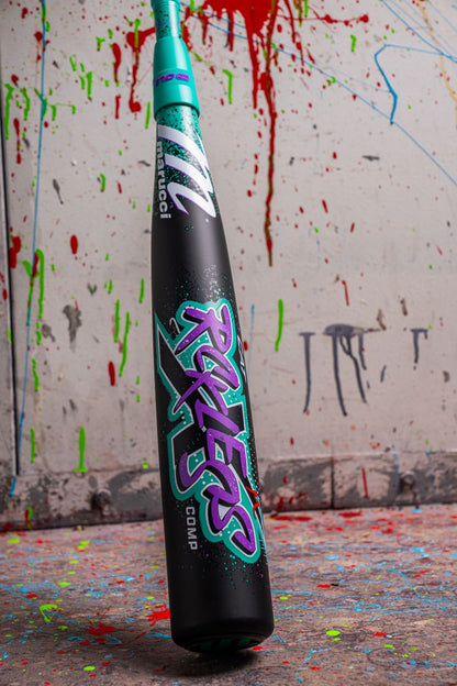 A black and teal Marucci 2026 CAT X RCKLESS Composite (-5) USSSA baseball bat (MSBCRC5 DEMO) stands upright on a paint-splattered floor, its bold graphics contrasting against a white wall streaked with red, green, and black paint.