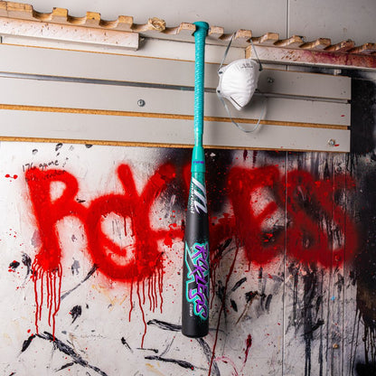 A 2026 Marucci CAT X RCKLESS Composite (-5) USSSA Baseball Bat and a white face mask hang on a paint-splattered wall, with "POGLESS" spray-painted in red graffiti behind them.