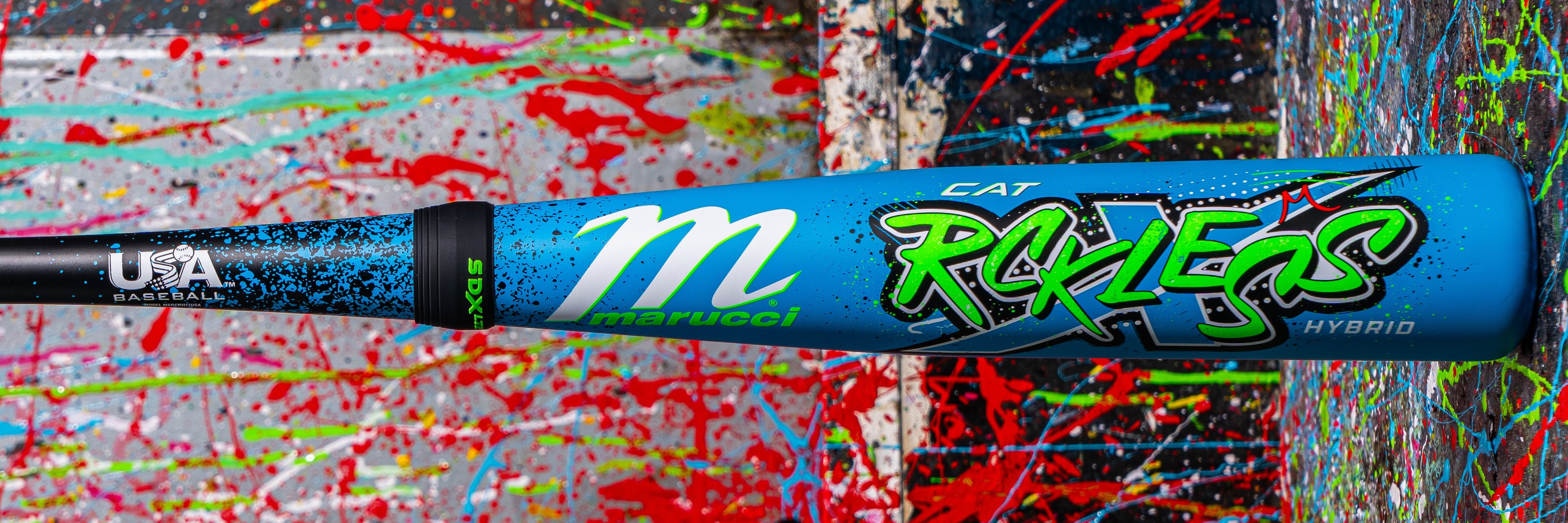 A blue Marucci CAT X baseball bat with vibrant green, white, and black graphics is displayed against a colorful, splatter-painted background. The words Reckless and Hybrid are visible on the barrel.