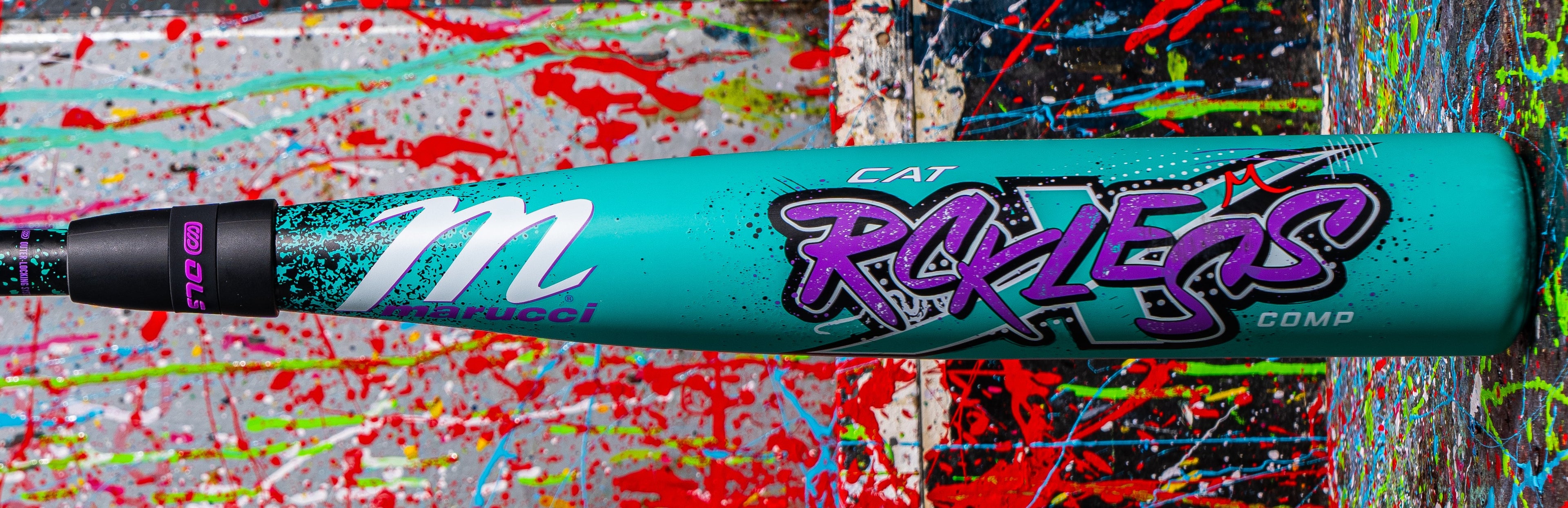 A turquoise Marucci CAT bat with bold, colorful RXT REKLSS COMP graphics rests on a splatter-painted background of red, blue, green, and yellow.