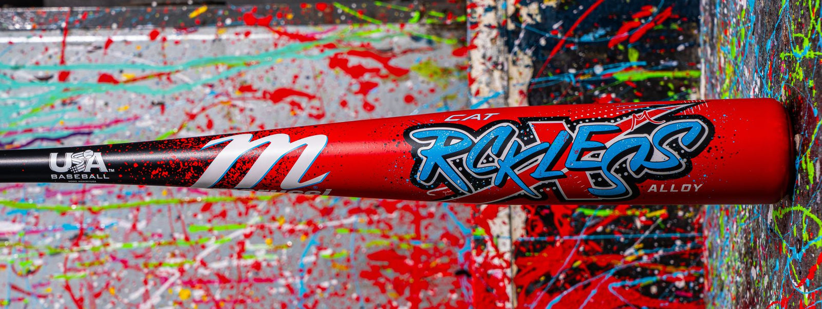 A red baseball bat with CAT REKLESS in blue graffiti-style lettering and USA Baseball logo, resting on a surface covered in colorful paint splatters.