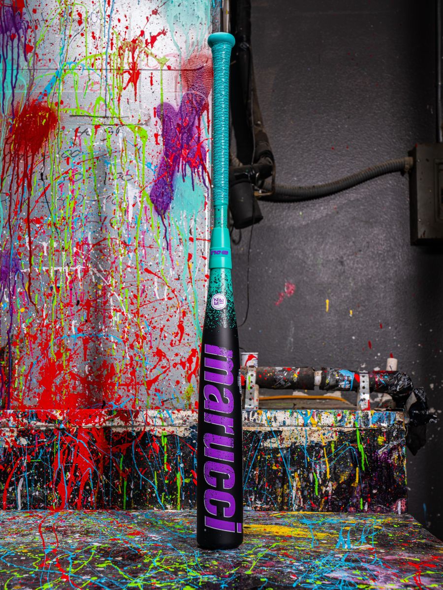A 2026 Marucci CAT X RCKLESS Composite (-5) 2 3/4" USSSA Baseball Bat (MSBCRC5 DEMO) stands upright in blue and pink against a splattered, colorful paint background with more paint on the floor and nearby objects.
