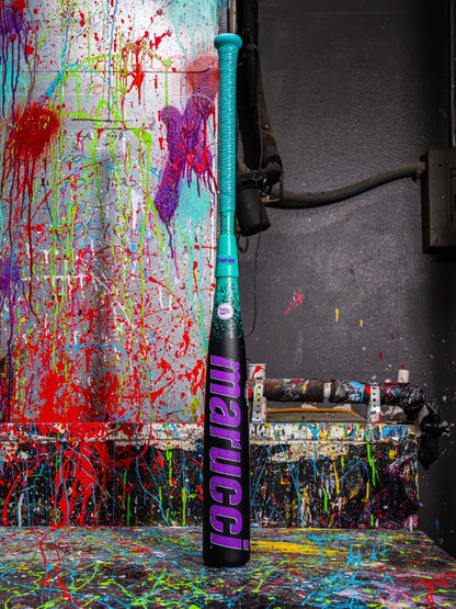 A 2026 Marucci CAT X RCKLESS Composite (-5) 2 3/4" USSSA Baseball Bat (MSBCRC5 DEMO) stands upright in blue and pink against a splattered, colorful paint background with more paint on the floor and nearby objects.