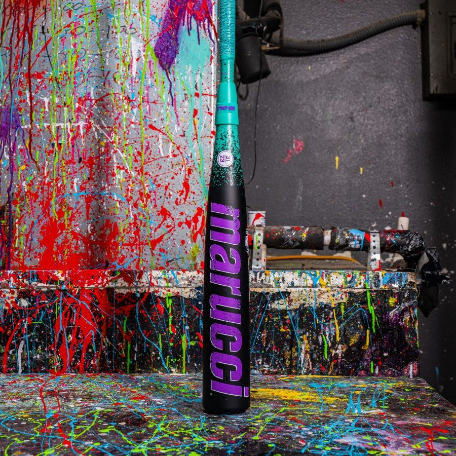 A black Marucci baseball bat with bright pink lettering stands upright on a splatter-painted surface, with a colorful, paint-splattered wall and shelf in the background.