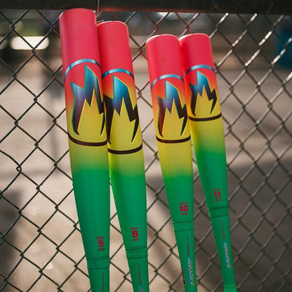 Four colorful USA certified bats with red, yellow, and green gradients plus flame graphics rest against a fence in bright sunlight. The standout is the 2026 Easton Hype Fire (-11) 2 5/8" USA Baseball Bat by Easton, with numbered handles.
