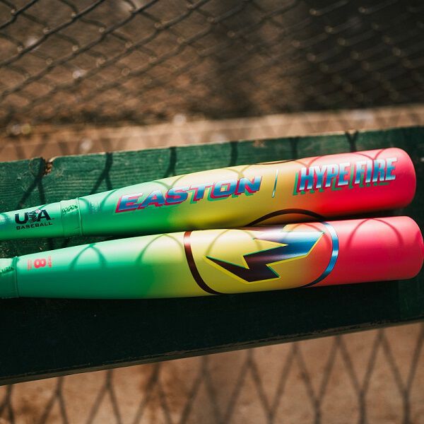 Two vibrant Easton Hype Fire (-11) 2 5/8" USA Baseball Bats (EUS6HYP11) with bold “EASTON” branding sit side by side on a green bench near a chain-link fence at the baseball field.