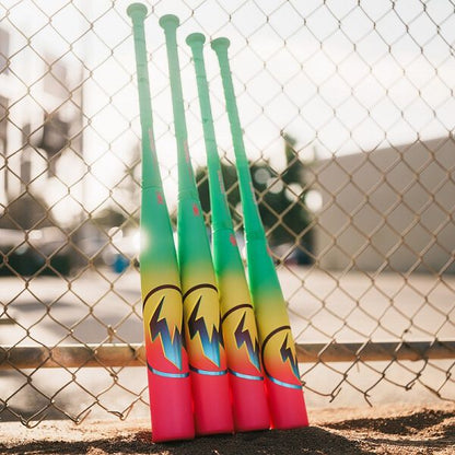 Four brightly colored baseball bats, including the 2026 Easton Hype Fire (-11) USA Baseball Bat by Easton with a green to yellow gradient and flame designs, stand upright against a chain-link fence on a sunny day.
