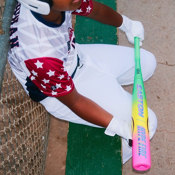 A young baseball player in a star jersey and white pants grips the Easton 2026 Hype Fire (-11) 2 5/8" USA Baseball Bat (EUS6HYP11), sitting on a green bench by a chain-link fence—perfect for youth players seeking top performance.