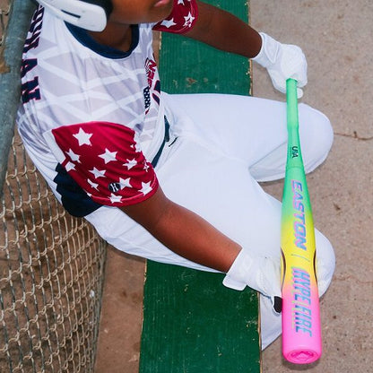 A young baseball player in a star jersey and white pants grips the Easton 2026 Hype Fire (-11) 2 5/8" USA Baseball Bat (EUS6HYP11), sitting on a green bench by a chain-link fence—perfect for youth players seeking top performance.
