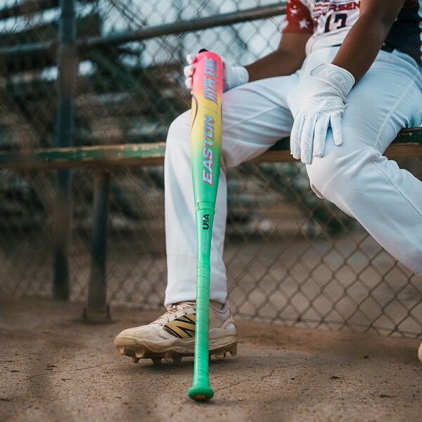A baseball player in white pants and gloves sits on a bench, holding the 2026 Easton Hype Fire (-11) 2 5/8" USA Baseball Bat (EUS6HYP11). The player’s face is hidden, with a chain link fence behind them.