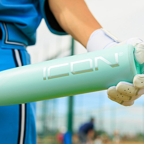 A person wearing blue athletic gear and white gloves holds a light green baseball bat with the word ICON written on it, standing on a sports field.