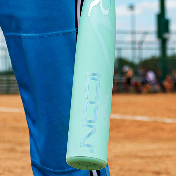 A person in blue athletic pants stands on a softball field, holding a mint green bat with the word ICON printed on it.