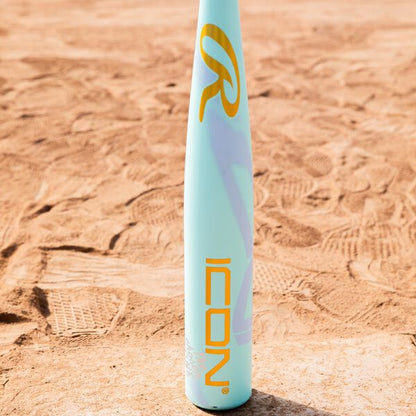 A 2026 Rawlings ICON CHOSEN ONE (-10) 2 5/8" USA Baseball Bat (RUS6IONE10) with yellow and purple graphics stands upright on a sandy field, showing its light blue color and visible shoe prints.