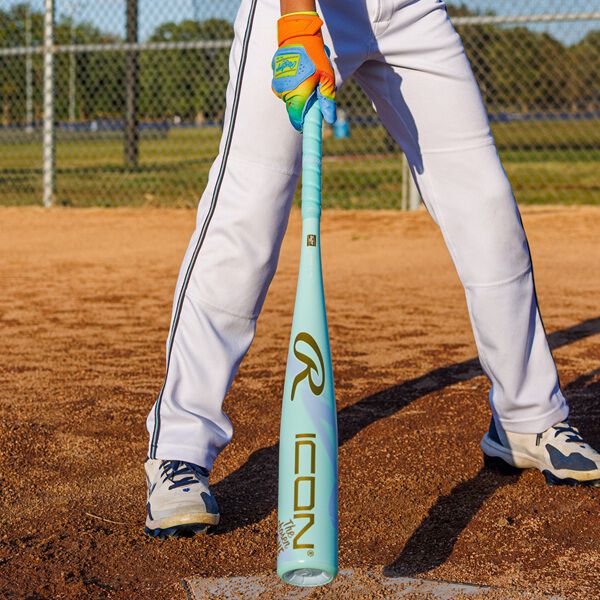 A baseball player stands at home plate holding a 2026 Rawlings ICON CHOSEN ONE (-10) 2 5/8" USA Baseball Bat: RUS6IONE10, wearing white pants, blue cleats, and orange batting gloves on a dirt field by a chain-link fence.