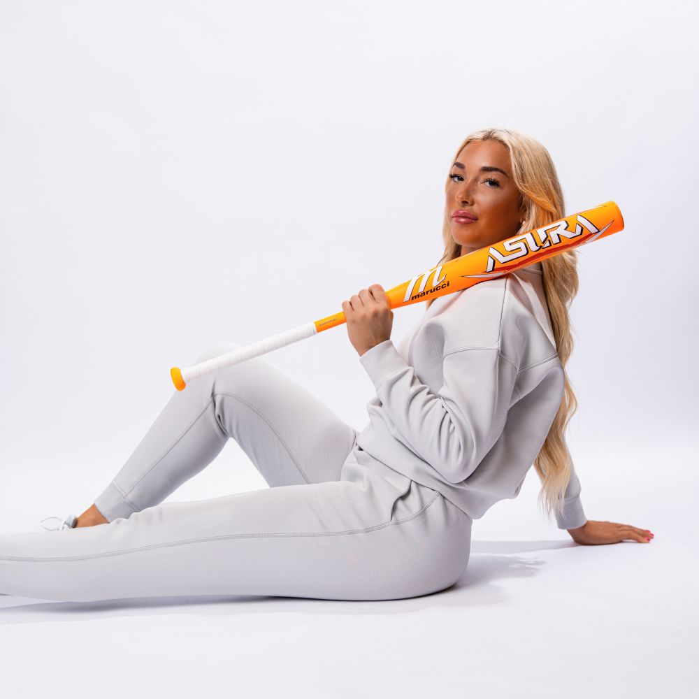 A person with long blonde hair in a light gray tracksuit sits with one knee bent, holding a Marucci 2025 ASURA Glow Citrus Spark (-11) Fastpitch Softball Bat (MFPAS11O) on their shoulder against a plain white background.