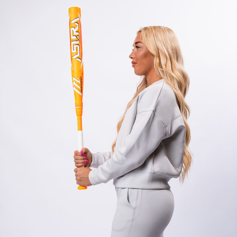 A woman with long blonde hair, in a light gray tracksuit, stands in profile holding a Marucci 2025 ASURA Glow Citrus Spark (-11) Fastpitch Softball Bat (MFPAS11O) upright with both hands against a plain white background.