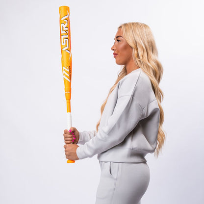 A woman with long blonde hair, in a light gray tracksuit, stands in profile holding a Marucci 2025 ASURA Glow Citrus Spark (-11) Fastpitch Softball Bat (MFPAS11O) upright with both hands against a plain white background.