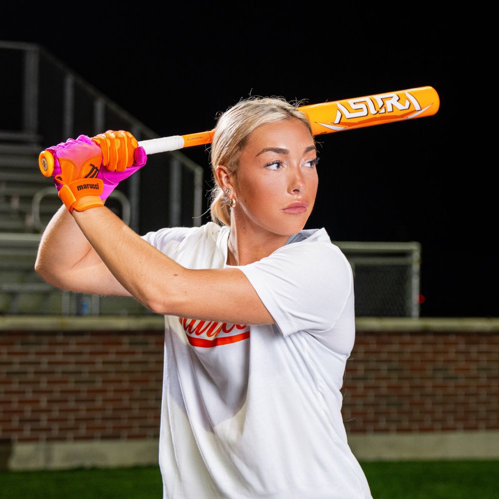 A blonde woman in a white shirt and neon gloves readies to swing a Marucci 2025 ASURA Glow Citrus Spark (-11) Fastpitch Bat (MFPAS11O) outdoors at night, with bleachers and a brick wall in the background.