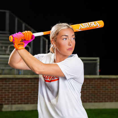 A blonde woman in a white shirt and neon gloves readies to swing a Marucci 2025 ASURA Glow Citrus Spark (-11) Fastpitch Bat (MFPAS11O) outdoors at night, with bleachers and a brick wall in the background.