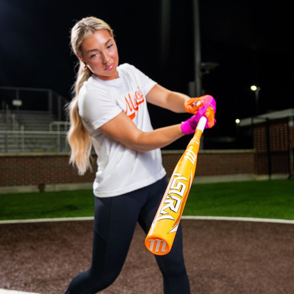 A young woman with long blonde hair wears a white shirt and pink gloves while swinging a Marucci 2025 ASURA Glow Citrus Spark (-11) Fastpitch Softball Bat (MFPAS11O) on a field at night, with stadium lights and empty bleachers behind her.