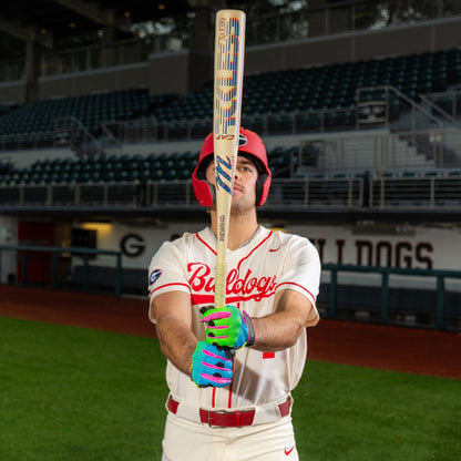 A baseball player in a white Bulldogs uniform and red helmet stands on the field, holding a Marucci 2026 CATX RCKLESS Rewind Hybrid (-3) BBCOR Baseball Bat (MCBCRHRC), with empty stadium seats visible in the background.