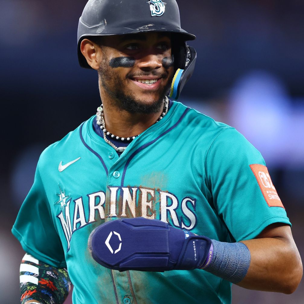 A baseball player in a teal Mariners jersey smiles on the field, wearing eye black, a beaded necklace, and an EvoShield Shorty Sliding Mitt 2.0 (WB57728) for hand protection. He stands ready with a blue mitt on his right hand; the background is blurred.