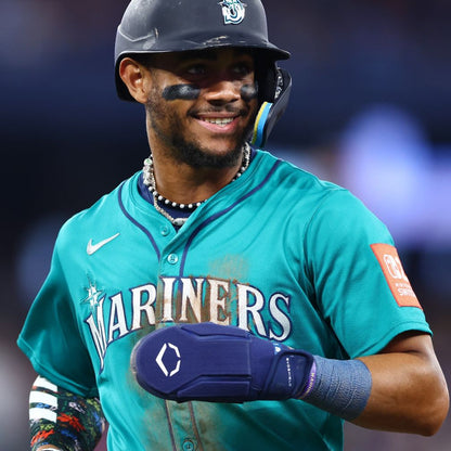A baseball player in a teal Mariners jersey smiles on the field, wearing eye black, a beaded necklace, and an EvoShield Shorty Sliding Mitt 2.0 (WB57728) for hand protection. He stands ready with a blue mitt on his right hand; the background is blurred.