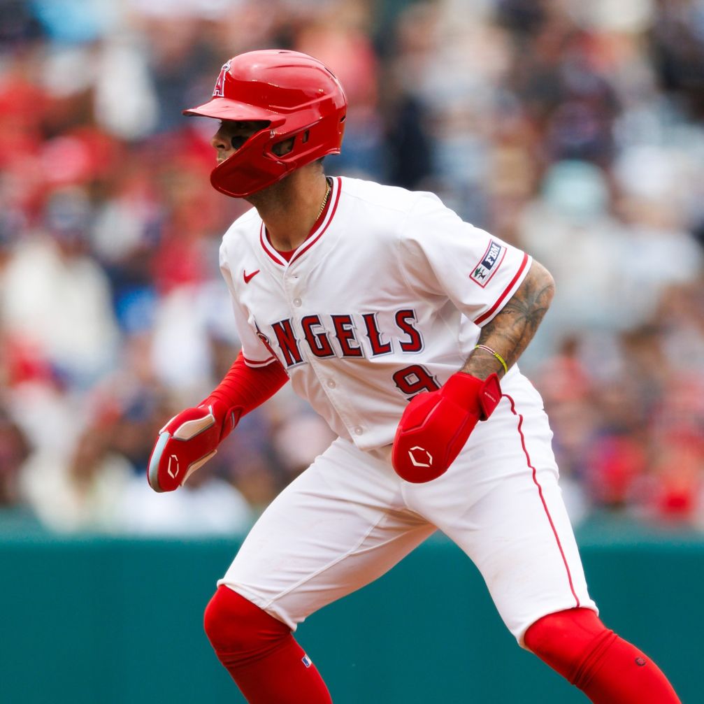 A baseball player in a white Los Angeles Angels uniform and red gloves stands ready on base, wearing an EvoShield Shorty Sliding Mitt 2.0 (WB57728) for added hand protection. The background shows blurred spectators as he looks to his right.