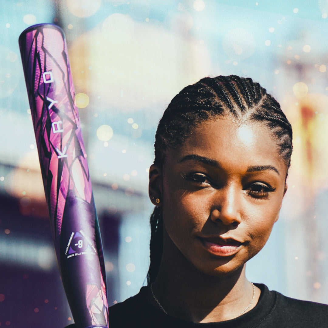A woman with braided hair confidently poses with a purple 2026 Louisville Slugger Kryo Thundernight Fastpitch Softball Bat over her shoulder, against a backdrop of vibrant, colorful bokeh lights.