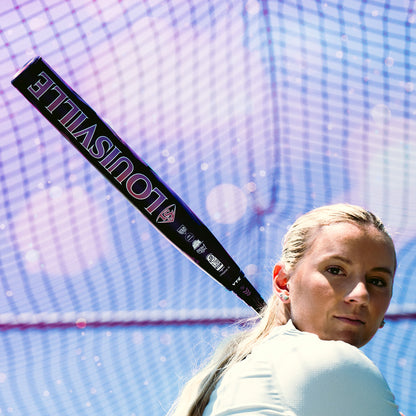 A woman prepares to swing the 2026 Louisville Slugger Kryo Thundernight Fastpitch Softball Bat, with blue netting and sunlight in the background.