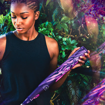 A woman in a sleeveless black top stands outdoors by greenery, looking down at a Louisville Slugger Kryo Thundernight Fastpitch Softball Bat in her hands as sunlight creates colorful light flares.