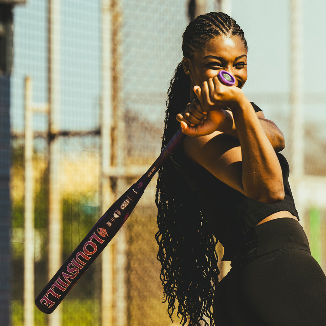 A woman with long braided hair smiles while holding a 2026 Louisville Slugger Kryo Thundernight Fastpitch Softball Bat, dressed in black and standing outdoors by a sunny chain-link fence.