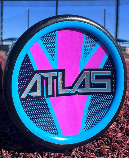 Close-up of a disc golf disc featuring a blue rim and pink-blue patterned center, boldly printed with "ATLAS"—inspired by the 2026 Louisville Slugger Atlas Hybrid (-3) BBCOR Baseball Bat: WBL4114010. Red turf, fence, and blue sky complete the scene.